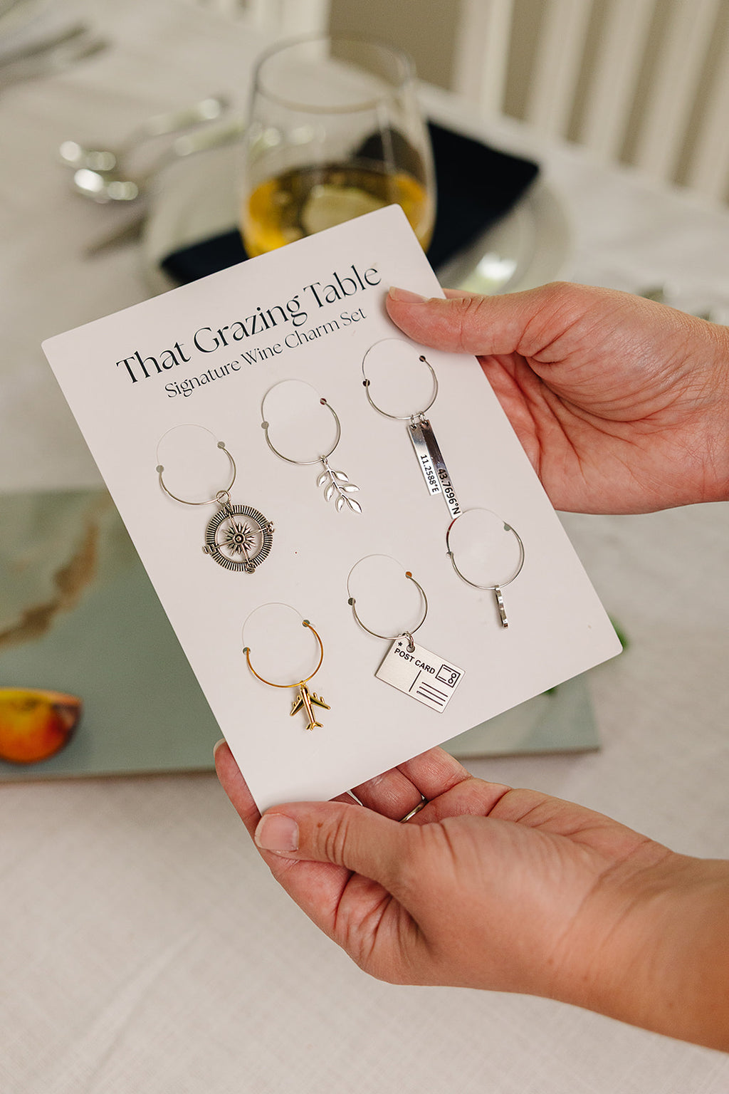 That Grazing Table Signature Wine Charm Set with six travel-inspired charms including compass, airplane, postcard, and olive branch on a branded keepsake card