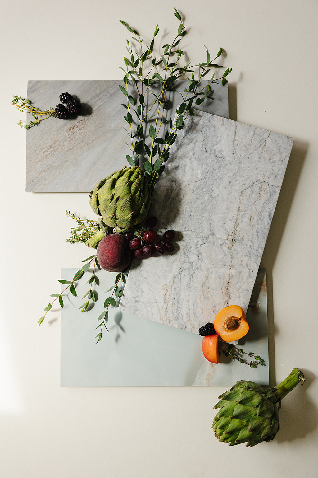 Handcrafted marble and Italian porcelain charcuterie boards by That Grazing Table, styled flat-lay with artichokes, stone fruit, grapes, and fresh greenery