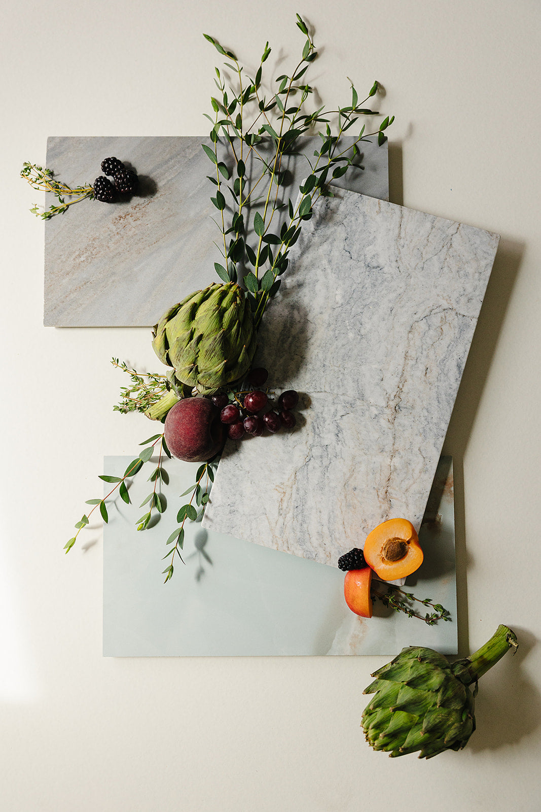 Handcrafted marble and Italian porcelain charcuterie boards by That Grazing Table, styled flat-lay with artichokes, stone fruit, grapes, and fresh greenery