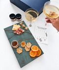 Sevilla Luxury Charcuterie Gift Set: olive green porcelain board with the three jams and butters, six wine charms (four on the card, two on glasses) and large navy gift box. Cheese, prosciutto, crackers, oranges, wine and glassware are styling only